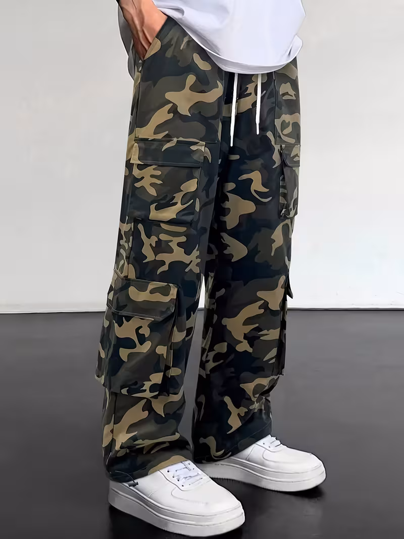 practical Men's Camouflage Print Multi-Pocket Cargo Pants, Elastic Waistband with Adjustable Legs, Fashion Wide-Leg Trousers 10 A studio photograph capturing a model from the waist down from a front three-quarter angle, wearing practical men's camouflage cargo pants in fern green. The relaxed, wide-leg trousers feature stacked side pockets with clean flap closures and white drawstrings hanging from an elastic waistband. Styled casually with crisp white platform sneakers on a smooth dark floor.