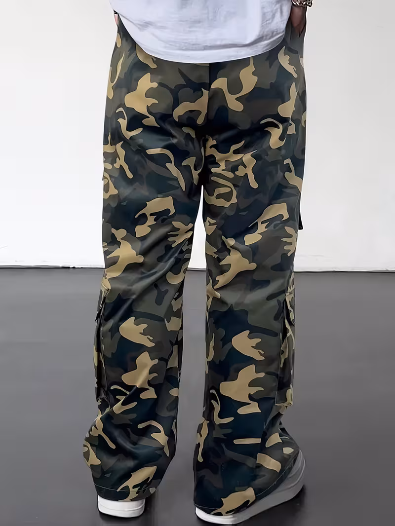practical Men's Camouflage Print Multi-Pocket Cargo Pants, Elastic Waistband with Adjustable Legs, Fashion Wide-Leg Trousers 11 A rear three-quarter studio shot of a model from the waist down showcasing the back profile of the men's fern green camouflage wide-leg cargo trousers. The structured material displays minimal wrinkling, hanging loosely over clean white sneakers on a dark floor against a clean white backdrop.