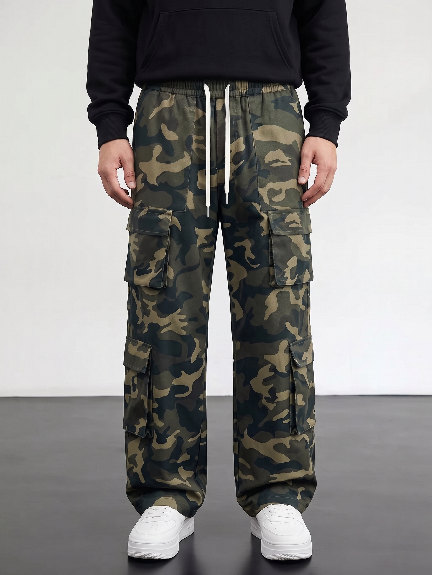 practical Men's Camouflage Print Multi-Pocket Cargo Pants, Elastic Waistband with Adjustable Legs, Fashion Wide-Leg Trousers 23 A full direct front-view studio photograph of a model wearing the fern green multi-pocket wide-leg camouflage cargo pants. The model is wearing a black crewneck sweatshirt and bright white sneakers. The symmetrical layout perfectly displays the dual upper utility pockets, mid-thigh cargo flaps, and the centered white drawstring.