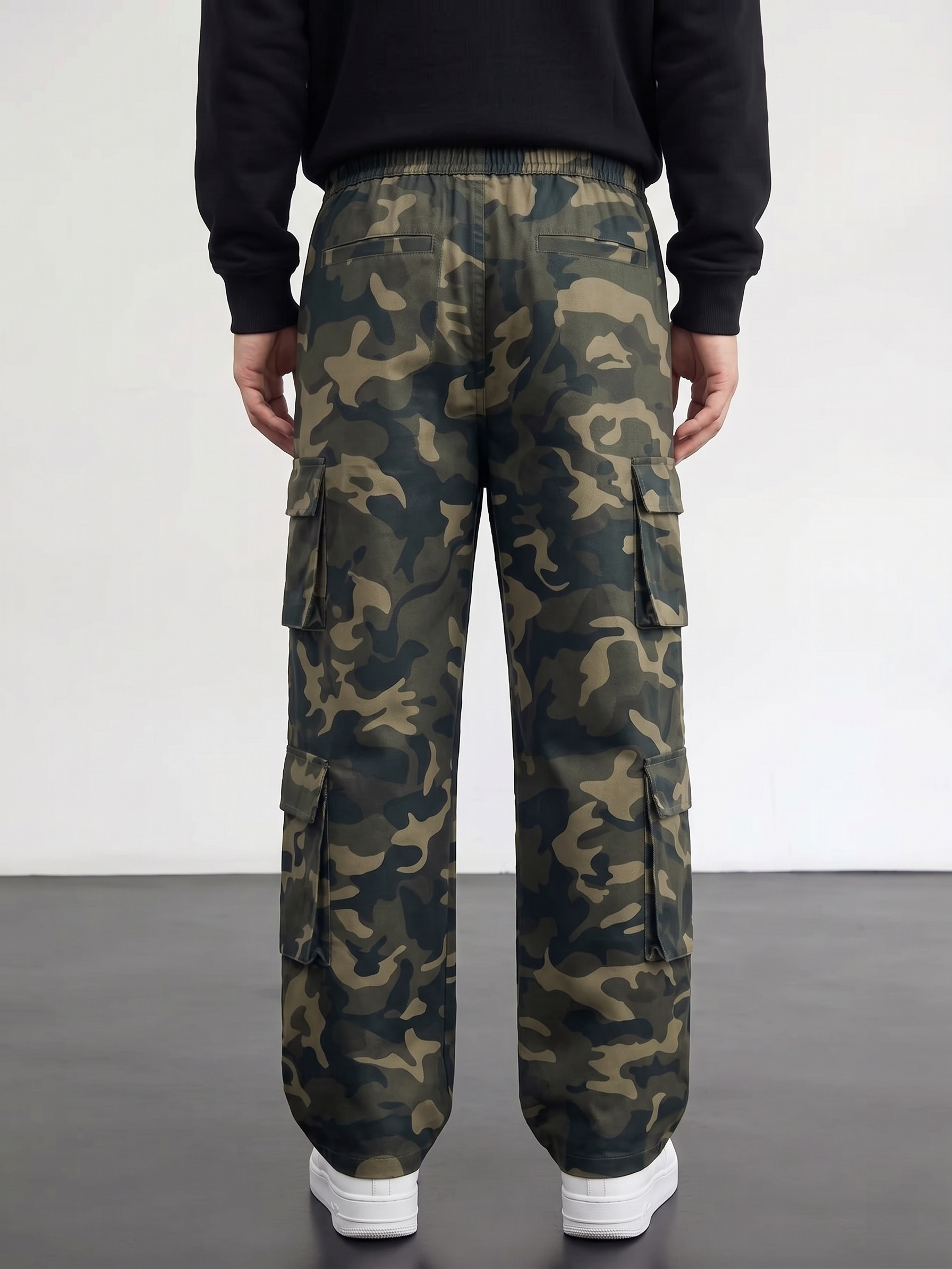 practical Men's Camouflage Print Multi-Pocket Cargo Pants, Elastic Waistband with Adjustable Legs, Fashion Wide-Leg Trousers 22 A direct rear-view studio photograph of a model from the waist down showcasing the backside of the fern green camouflage cargo pants. The model wears a black sweatshirt and white sneakers. The view highlights clean welt-style rear back pockets, the gathered elastic waistband, and the wide leg openings.