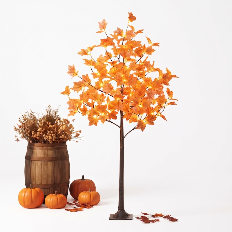 A full, eye-level studio photograph of the 1 pc 6ft/180cm Artificial Lighted Maple Tree, fully illuminated with warm white lights, standing against a seamless white background. The tree's brown trunk is set in its square base, surrounded by a rustic wooden barrel planter, three artificial pumpkins, and scattered maple leaves on the white surface. The warm glow from the leaves creates a cozy ambiance.