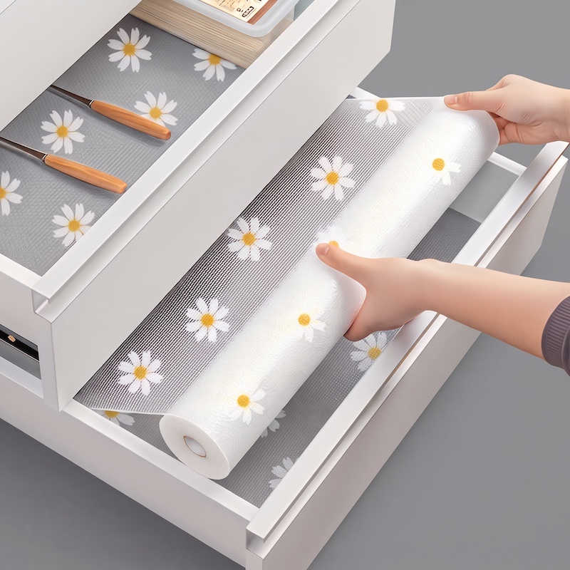 Hands unroll a textured grey cabinet liner 1 roll of daisy design into a white kitchen drawer, with another lined drawer visible above.