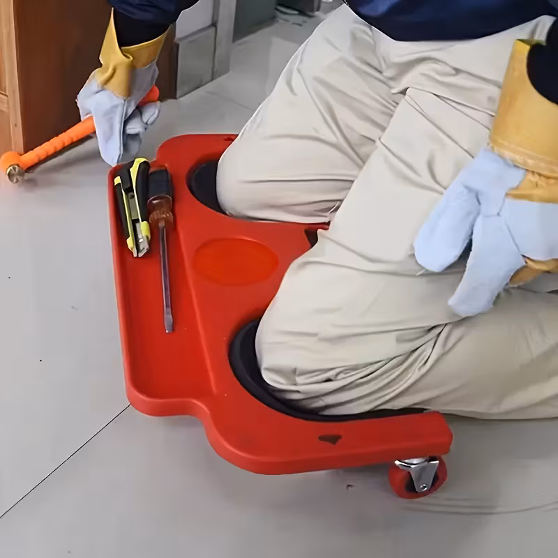 A Rolling Knee Board Platform with Wheels, Made of ABS Material, Featuring a Multifunctional Built-in Tool Tray and a Comfortable Cushion. Suitable for Mechanics, Carpenters, Flooring, and Automotive Repairs. Valentine's Day Gifts for Men 19 A person wearing tan pants, a blue jacket, and large white and yellow gloves is kneeling on a red Rolling Knee Board Platform with Wheels. The top section of the platform has black padded contours for the knees. Integrated tool storage compartments on the right hold a utility knife and a screwdriver. One small red swivel caster wheel is clearly visible on the bottom-right corner. The person’s left hand holds an orange mallet on the light grey floor.