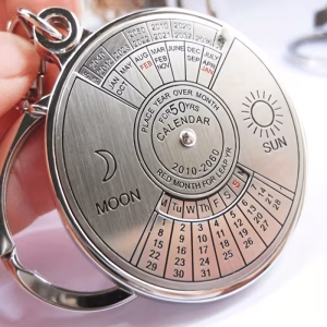 A professional high-definition macro photograph of the Calendar Keychain Retro being held by a user. The hardware is constructed from brushed and polished silver-toned metal, featuring a multi-tier rotating dial. The upper section displays years from 2010 to 2060, while the middle section lists months, with "JAN" and "FEB" highlighted in red for leap year alignment. The lower grid provides a day-to-date matrix (M through S). Engraved central text reads "PLACE YEAR OVER MONTH FOR 50 YRS CALENDAR." The chassis is adorned with celestial "MOON" and "SUN" iconographies and is attached to a heavy-duty split-ring connector. The focus is sharp on the laser-etched typography and the mechanical pivot point.