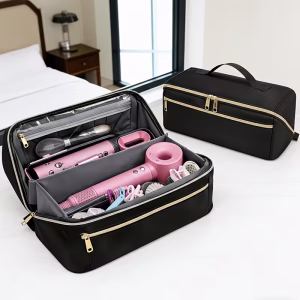Two black color clutch bag hot hair tools travel bags are resting on a white desk in a bedroom setting. One travel bag is open, showing a neat interior with pink hair tools like a blow dryer and flat iron, plus various hair accessories and makeup brushes. The other black color clutch bag hot hair tools travel bag is closed, sitting behind it to display its sleek design and gold zipper accents. A bed and nightstand are visible in the background.